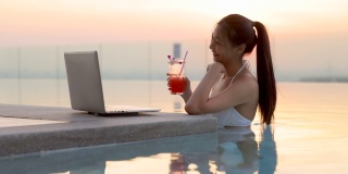穿着比基尼的亚洲女人在泳池喝鸡尾酒，在笔记本电脑上搜索电视剧，在互联网流媒体服务上选择电影休闲在早上，晚上。模糊的背景光线，日落，日出