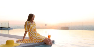 轻松放松的亚洲女商人生活方式在美丽的夏季连衣裙坐在池边，享受清爽的鸡尾酒在一个杯子与冰在模糊的背景美丽的日落天空。