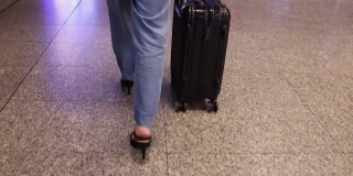 女人穿着牛仔裤长裤，黑色鞋跟鞋，带着黑色行李箱沿着机场航站楼走着，看腿的底部。