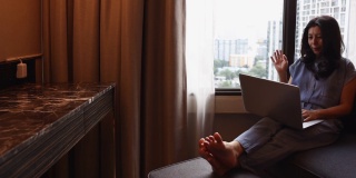 亚洲妇女视频电话笔记本电脑，而坐在现代椅子在酒店房间商务旅行。有魅力的女商人与家人进行远距离交流。