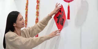 快乐的亚洲妇女在准备中国新年期间贴在墙上的汉字“财富”春联
