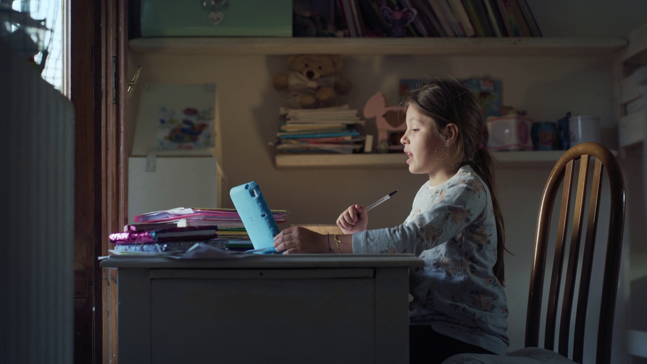 图为，一名小女孩在家中的远程教育教室里，与老师一起用平板电脑进行在线授课，并在书桌上用笔记本书写的真实画面。Covid