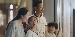 亚裔家庭的父母和孩子站在商场的橱窗里看