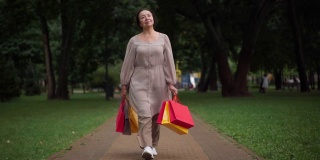 宽镜头的肖像华丽自信的资深妇女步行与购物袋在公园微笑。美丽优雅的白种人退休老人满足购物在户外散步在春天夏天的一天。