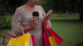 一位无法辨认的白人老年妇女拿着购物袋站在那里，用智能手机在线发短信，面带微笑。积极自信的女士拿着保温瓶在户外上网。视频素材模板下载