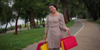 自信美丽的老女人站在公园的小巷微笑着看着镜头，肩上挂着购物袋。积极的白人优雅的女士摆姿势与购买户外的肖像。