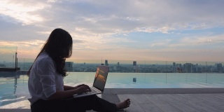 年轻女子在工作，用笔记本电脑坐在摩天大楼的泳池旁。自由职业，假期，远程工作，社会距离，电子学习，在线会议的概念。本空间