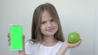 小女孩用绿色屏幕展示苹果和手机模拟手机、手机、手机。绿屏智能手机的色度键用于广告。水果、健康饮食、儿童口腔保健视频素材模板下载