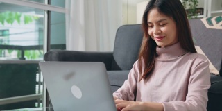 年轻快乐的女人坐在家里的沙发上，用笔记本电脑在键盘上打字。