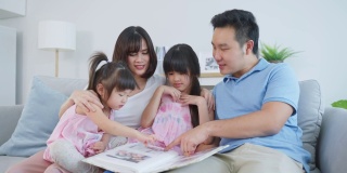 亚洲家庭看着旧相册上的照片回忆过去。年轻漂亮的夫妇和年幼的女儿坐在一起，翻动着相册，享受着回忆彼此关系的时光。