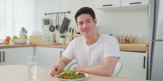 亚洲活跃的帅哥拒绝吃甜甜圈，喜欢吃健康食品。年轻有魅力的男性，在厨房的碗里吃蔬菜沙拉，在桌子上喝干净的水。饮食与健康理念