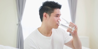亚洲活跃的强壮男人的肖像在卧室里拿着干净的水。吸引人的口渴男性饮料或喝一小口在杯子的天然矿物质，为了健康和幸福醒来后看着相机。