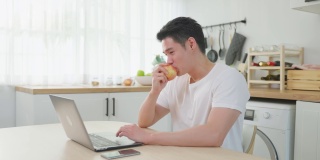亚洲商人在家工作时吃苹果健康食品。有吸引力的英俊年轻男性感觉快乐和享受，使用笔记本电脑学习在线学习和吃水果在家里的厨房。