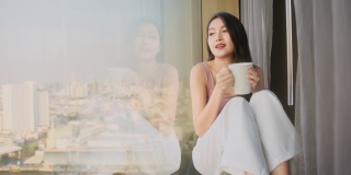 幸福迷人的亚洲女性放松休闲坐在窗边看外面手握热饮料在酒店房间