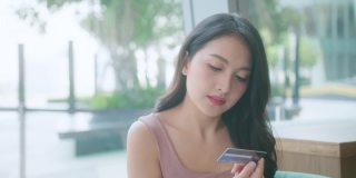 美丽的亚洲年轻女性与欢快的表情，持有智能手机和信用卡，银行在线，快乐的年轻女孩持有塑料信用卡，而使用手机在咖啡馆