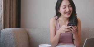 聊天成瘾的亚洲女性幸福快乐，在家里用智能手机短信与朋友交流，在沙发上开心微笑