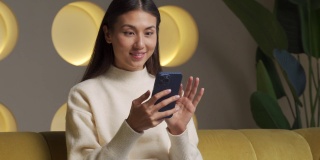 可爱的年轻亚洲女子坐在客厅的沙发上，用智能手机在她的手中进行网上购物