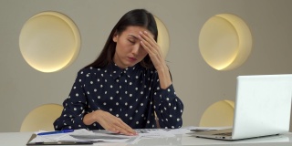 一名亚洲女性在办公室用手捂住脸，在办公桌上用笔记本电脑工作后，心情烦躁