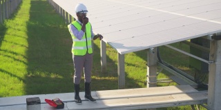 在太阳能发电厂安装太阳能电池板的过程中，非洲的年轻工程师穿着防护服和反光布爬上并站在结构上，通过无线电与团队联系