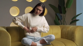 一名年轻的亚裔女性，穿着运动衫，双手摸着肚子，坐在家里的沙发上感到腹部疼痛视频素材模板下载