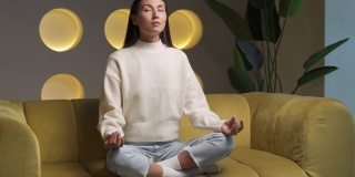 该上瑜伽课了。有吸引力的年轻亚洲妇女训练和坐在一个瑜伽莲花姿势在沙发上