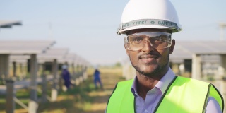 特写，肖像:在太阳能农场，非洲年轻工程师穿着防护服，戴着安全眼镜和头盔，开心地看着镜头