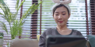 年轻迷人的亚洲女性在家里工作，手用平板电脑，检查业务数据，图表，计划报告，休闲放松在沙发上，在客厅居家隔离的概念