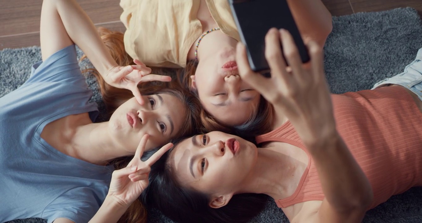 上图是一群亚洲女士在家中客厅的地板上躺在地毯上，拿着智能手机微笑着拍下回忆的照片。生活方式活动隔离概念。