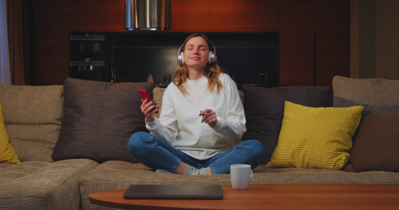 快乐的年轻女性戴着无线耳机躺在沙发上用手机app听音乐缓解压力