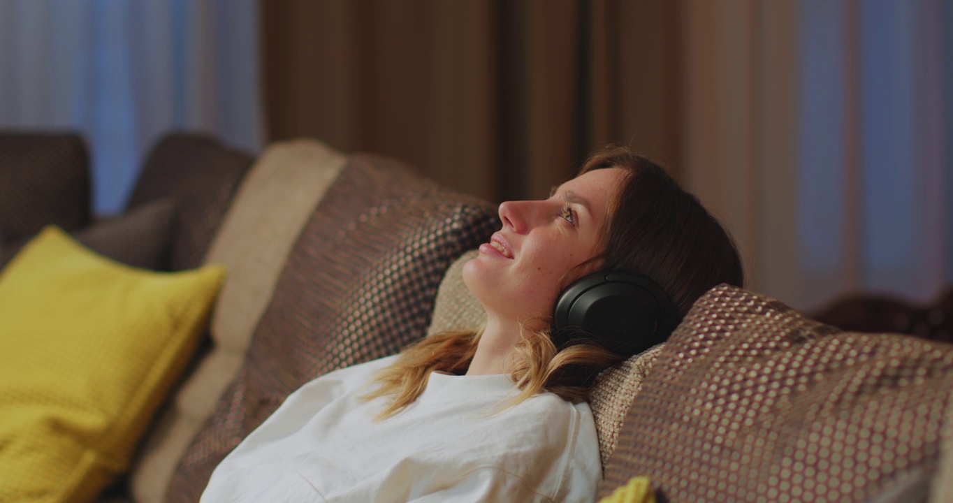 冷静的年轻女子戴上现代无线耳机，放松在舒适的沙发上，听着音乐，无压力的概念