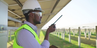 侧视图:非洲青年工程师，穿着防护服，戴着安全眼镜和安全帽，通过无线电与团队联系