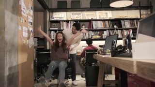 两个亚洲同事在办公室里通过滑动办公椅开玩笑。视频素材模板下载