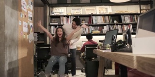 两个亚洲同事在办公室里通过滑动办公椅开玩笑。