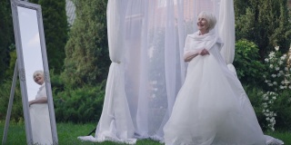 广角镜头快乐的高级白人新娘在白色优雅的婚纱纺纱在婚礼祭坛户外欣赏镜子的反射。快乐自信的女人在春天夏天公园结婚的肖像。