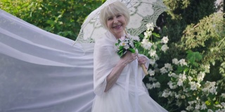 积极自信的老年女性穿着婚纱，戴着面纱，打着遮阳伞，在春天夏天的公园里摆出慢动作。快乐的白人美丽的新娘看着镜头微笑的肖像。