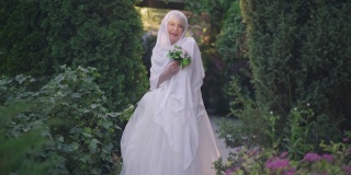 宽镜头快乐美丽自信的年长新娘在婚礼礼服和白色披肩在阳光下在户外摆姿势。美丽微笑的白人退休人员站在阳光微笑。缓慢的运动。