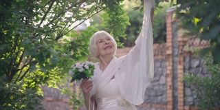 微笑着，白发的老新娘站在阳光下，用慢镜头举起披巾的手。自信自豪的白人妇女在阳光下户外婚礼的肖像。