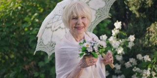 优雅的老年新娘捧着花束和遮阳伞，在阳光明媚的春夏季的公园里摆出慢动作。美丽快乐的白种人女人的肖像与灰色的头发看着镜头微笑。