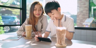一个男孩在咖啡厅里用智能手机玩游戏，旁边站着一位女商人。女人和孩子说话。幸福和微笑。桌上有冰咖啡和巧克力。教育与科技理念