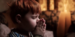 一个小男孩正在一间黑暗的房间里祈祷。一个七到十岁的红头发男孩合掌祈祷。