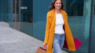 女商人提着购物袋，穿着黄色外套，走在户外社团建筑的背景下视频素材模板下载
