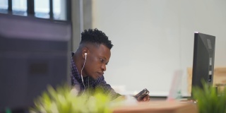 年轻的非洲裔美国人戴着耳机，手持智能手机，坐在办公室里发短信，使用应用程序在社交媒体上聊天，在手机上上网。新一代的现代生活方式。