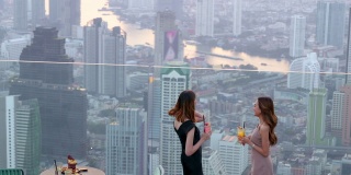 4K亚洲女性朋友在夏日夕阳下，在摩天大楼的屋顶餐厅一起喝鸡尾酒聊天。