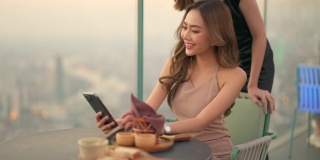 日落时分，4K的亚洲女性朋友在摩天大楼的屋顶餐厅一起用智能手机自拍。