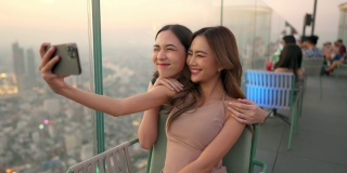 日落时分，4K的亚洲女性朋友在摩天大楼的屋顶餐厅一起用智能手机自拍。
