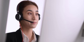 亚洲女性呼叫中心工作时使用耳机看电脑屏幕。肖像微笑的亚洲女性客户服务代理戴着耳机工作在办公室的背景