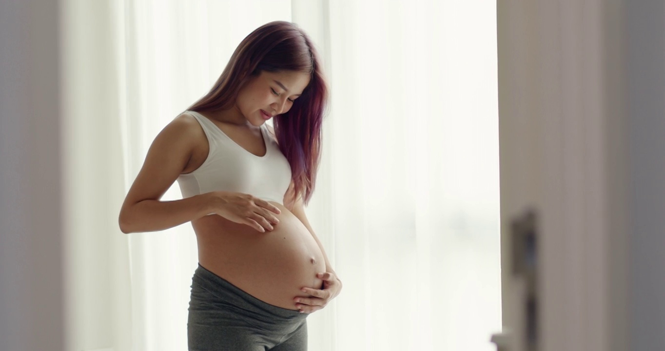 一位美丽的亚洲孕妇站在窗前，抚摸着自己的肚子。