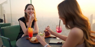4K亚洲女性朋友在夏日夕阳下在摩天大楼的屋顶餐厅共进晚餐。