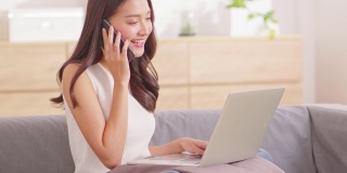 美丽的亚洲妇女穿着休闲毛衣工作在笔记本电脑上微笑，并与客户在智能手机交谈，以获得成功的工作在家里的想法要求。自由职业妇女工作在线销售营销