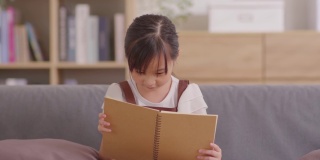可爱的蹒跚学步的小女孩在家里的沙发上看书。快乐学前儿童读物，享受自学教育。教育的概念